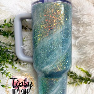 May include: A large, glitter-covered tumbler with a handle. The tumbler features a gradient of teal and blue with white swirls, and a clear lid. The handle and lid are light gray. The tumbler has the text "Tipsy Tinkerbell Tumblers & More".