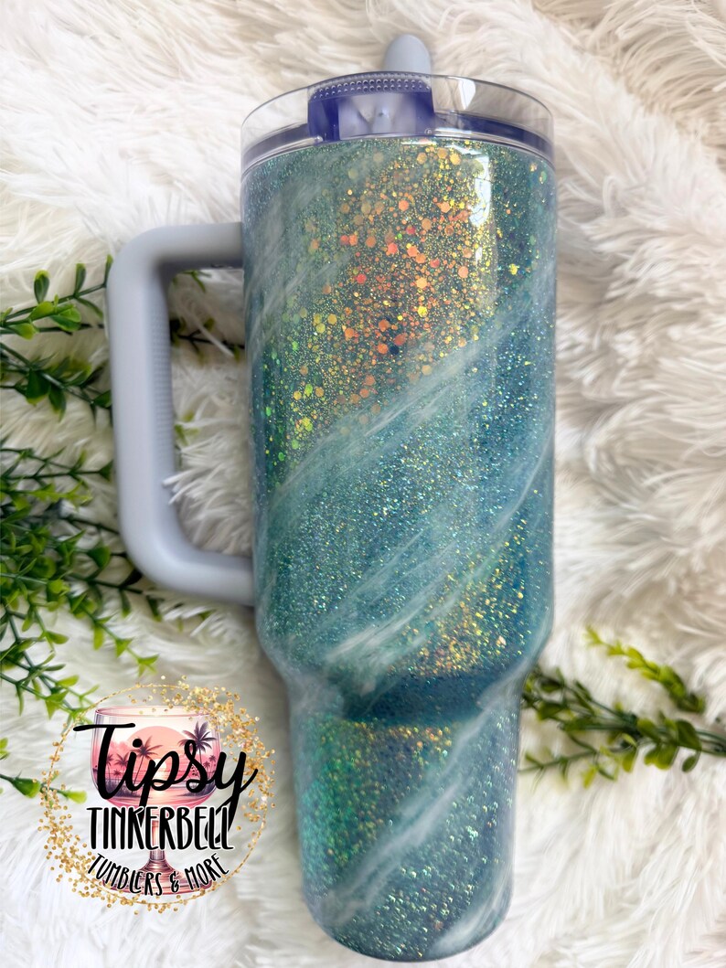 May include: A large, glitter-covered tumbler with a handle. The tumbler features a gradient of teal and blue with white swirls, and a clear lid. The handle and lid are light gray. The tumbler has the text "Tipsy Tinkerbell Tumblers & More".