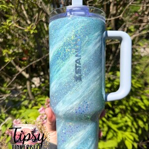 May include: A blue and white glitter Stanley tumbler with a handle. The tumbler has a light blue lid and handle. The word "Stanley" is printed on the side. The tumbler is decorated with a swirling pattern of blue and white glitter.