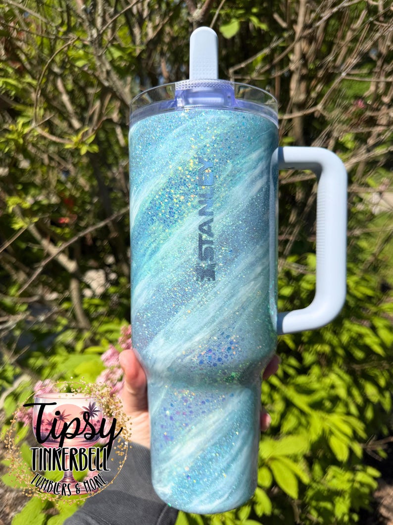 May include: A blue and white glitter Stanley tumbler with a handle. The tumbler has a light blue lid and handle. The word "Stanley" is printed on the side. The tumbler is decorated with a swirling pattern of blue and white glitter.