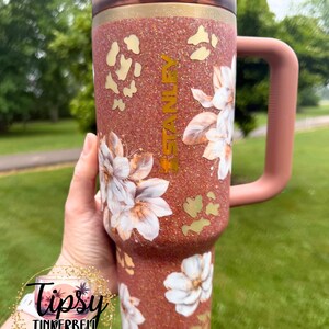 May include: A copper-coloured glitter tumbler with a handle and a dark lid. The tumbler features white floral decals, gold leopard print accents, and the word STANLEY printed vertically in gold. A circular logo with the text Tipsy Tinkerbell Tumblers & More is visible.