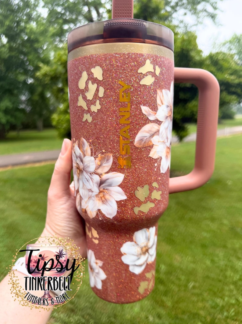 May include: A copper-coloured glitter tumbler with a handle and a dark lid. The tumbler features white floral decals, gold leopard print accents, and the word STANLEY printed vertically in gold. A circular logo with the text Tipsy Tinkerbell Tumblers & More is visible.