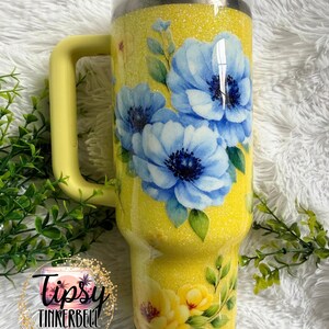 May include: A yellow glitter tumbler with a handle, featuring a floral design of blue and yellow flowers with green leaves. The tumbler has a white lid and a yellow handle. The text "Tipsy Tinkerbell Tumblers & More" is visible.