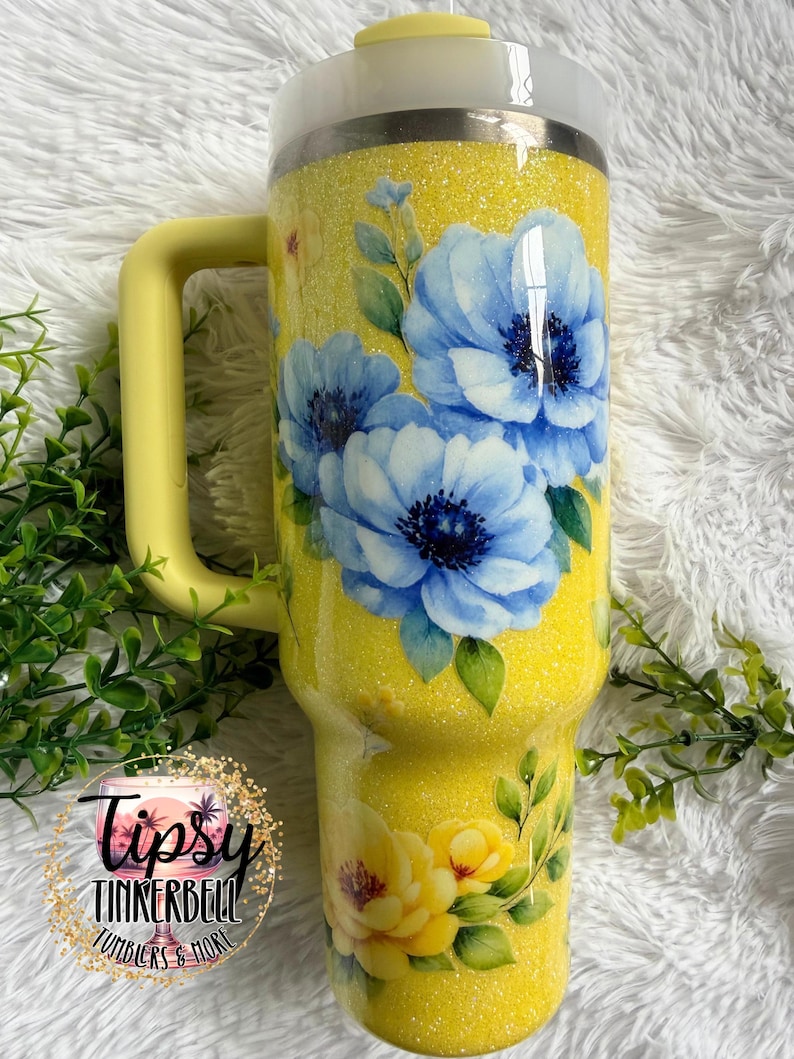 May include: A yellow glitter tumbler with a handle, featuring a floral design of blue and yellow flowers with green leaves. The tumbler has a white lid and a yellow handle. The text "Tipsy Tinkerbell Tumblers & More" is visible.