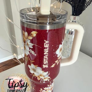 Puede incluir: Un vaso Stanley rojo brillante con un dise&ntilde;o floral. El vaso tiene un asa blanca y una tapa transparente. La palabra "STANLEY" est&aacute; impresa en el lateral. El vaso est&aacute; decorado con flores blancas y doradas.