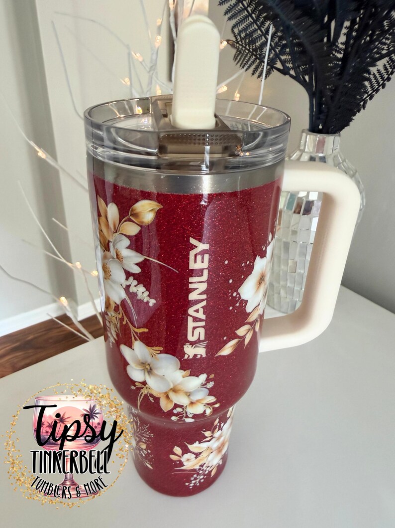 Puede incluir: Un vaso Stanley rojo brillante con un dise&ntilde;o floral. El vaso tiene un asa blanca y una tapa transparente. La palabra "STANLEY" est&aacute; impresa en el lateral. El vaso est&aacute; decorado con flores blancas y doradas.