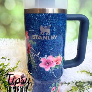 Może przedstawiać: Niebieski, brokatowy kubek Stanley z ciemnoniebieskim uchwytem i pokrywką. Kubek ozdobiony jest kwiatowym wzorem z r&oacute;żowymi kwiatami hibiskusa i zielonymi liśćmi. Logo Stanley jest wygrawerowane z przodu. Słomka jest umieszczona w środku pokrywki.