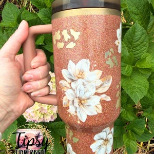 May include: A copper-coloured glitter tumbler with a handle, featuring white floral decals and gold-toned leopard print accents. The cup has a dark translucent lid with a straw opening and a solid copper-coloured handle attached to the side.