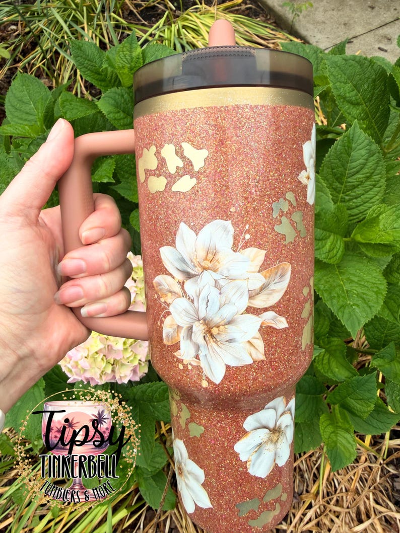May include: A copper-coloured glitter tumbler with a handle, featuring white floral decals and gold-toned leopard print accents. The cup has a dark translucent lid with a straw opening and a solid copper-coloured handle attached to the side.