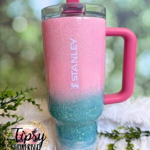 May include: A Stanley tumbler featuring a pink glitter top half and a teal glitter bottom half that transitions into a white wave pattern at the base. The cup includes a pink handle, a clear lid, and a pink straw, with the Stanley logo in white.