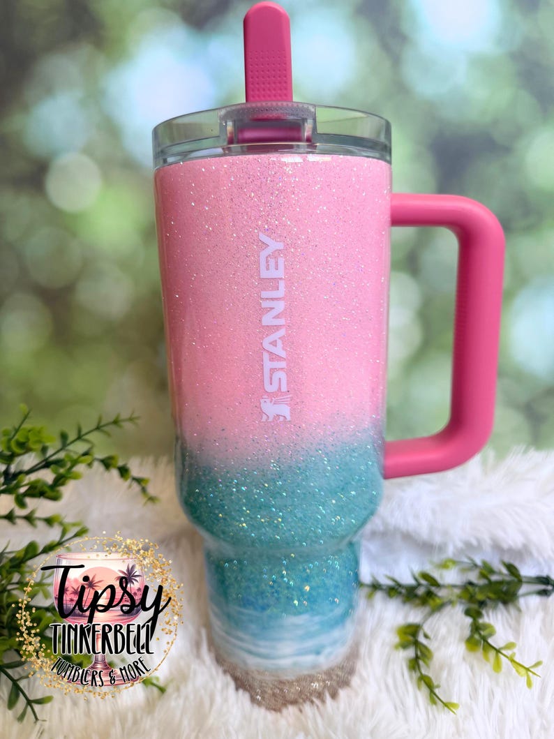 May include: A Stanley tumbler featuring a pink glitter top half and a teal glitter bottom half that transitions into a white wave pattern at the base. The cup includes a pink handle, a clear lid, and a pink straw, with the Stanley logo in white.