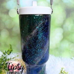 May include: A cylindrical tumbler with a white lid and handle, featuring a dark blue and emerald green glitter finish with swirling silver accents. The surface is glossy and textured with embedded reflective particles throughout the design.