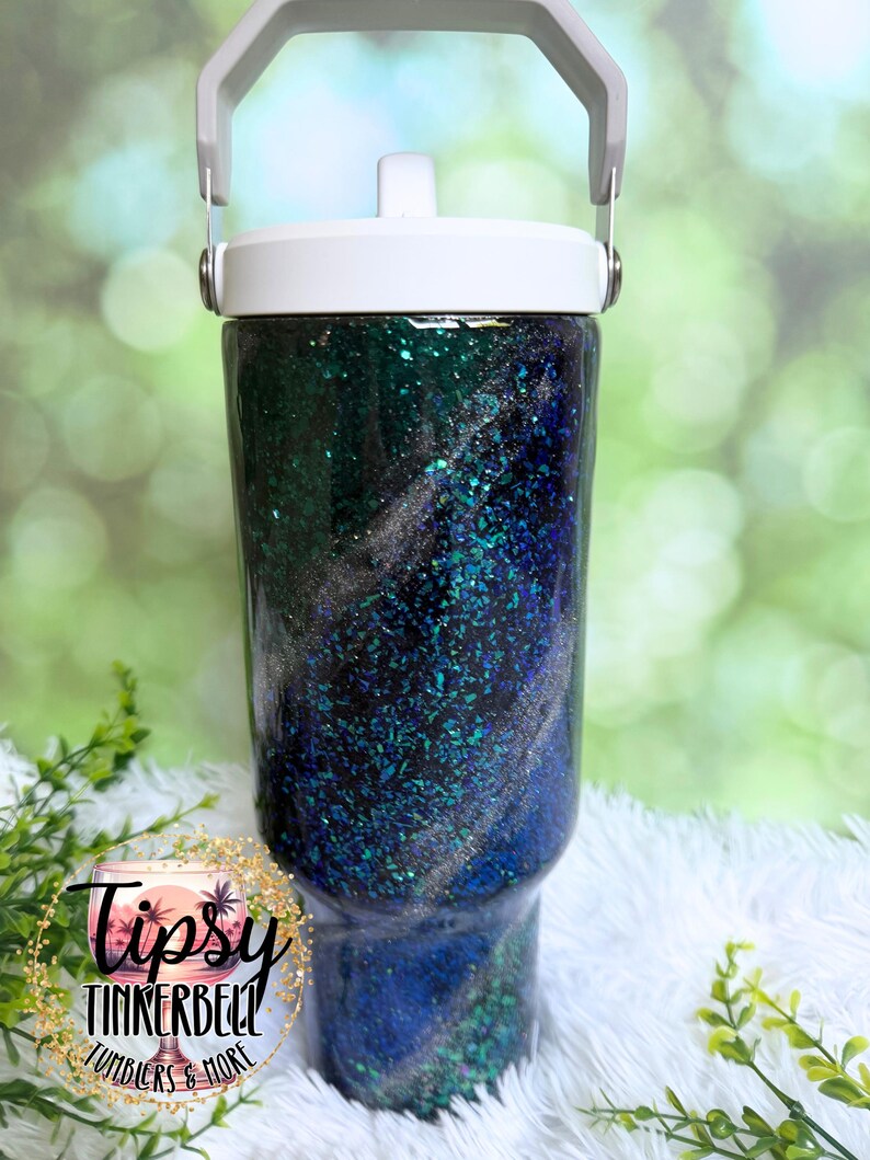 May include: A cylindrical tumbler with a white lid and handle, featuring a dark blue and emerald green glitter finish with swirling silver accents. The surface is glossy and textured with embedded reflective particles throughout the design.