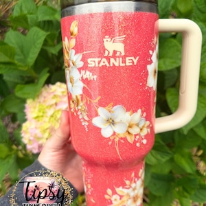 K&ouml;nnte beinhalten: Ein korallenfarbener Stanley-Becher mit Glitzer-Finish, verziert mit einem wei&szlig;en Blumenmuster und einem cremefarbenen Henkel. Der Becher hat einen dunkel durchscheinenden Deckel und das Stanley-Logo in Wei&szlig;. Ein kreisf&ouml;rmiges Logo mit dem Text Tipsy Tinkerbell Tumblers & More ist in der unteren linken Ecke zu sehen.