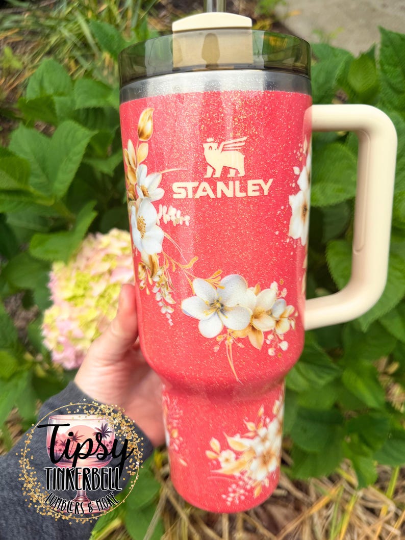 K&ouml;nnte beinhalten: Ein korallenfarbener Stanley-Becher mit Glitzer-Finish, verziert mit einem wei&szlig;en Blumenmuster und einem cremefarbenen Henkel. Der Becher hat einen dunkel durchscheinenden Deckel und das Stanley-Logo in Wei&szlig;. Ein kreisf&ouml;rmiges Logo mit dem Text Tipsy Tinkerbell Tumblers & More ist in der unteren linken Ecke zu sehen.
