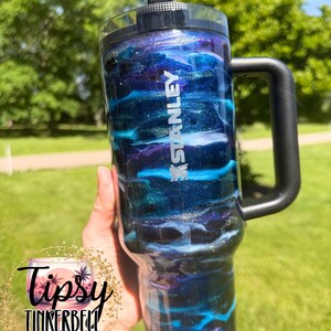 May include: A Stanley tumbler with a black handle and lid. The tumbler features a swirling design of blue, purple, and silver glitter, with the Stanley logo in white. The tumbler is held against a background of green grass and trees.
