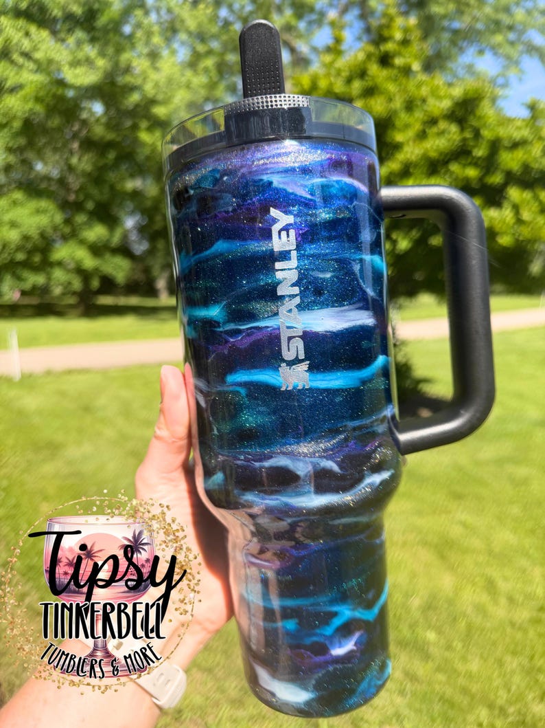 May include: A Stanley tumbler with a black handle and lid. The tumbler features a swirling design of blue, purple, and silver glitter, with the Stanley logo in white. The tumbler is held against a background of green grass and trees.
