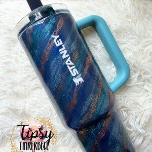Puede incluir: Un vaso Stanley con un dise&ntilde;o de purpurina azul, verde azulado y dorado. El vaso tiene un asa azul claro y el logotipo de Stanley en blanco. Tiene una tapa azul oscuro y una pajita negra. Un recipiente para bebidas elegante.