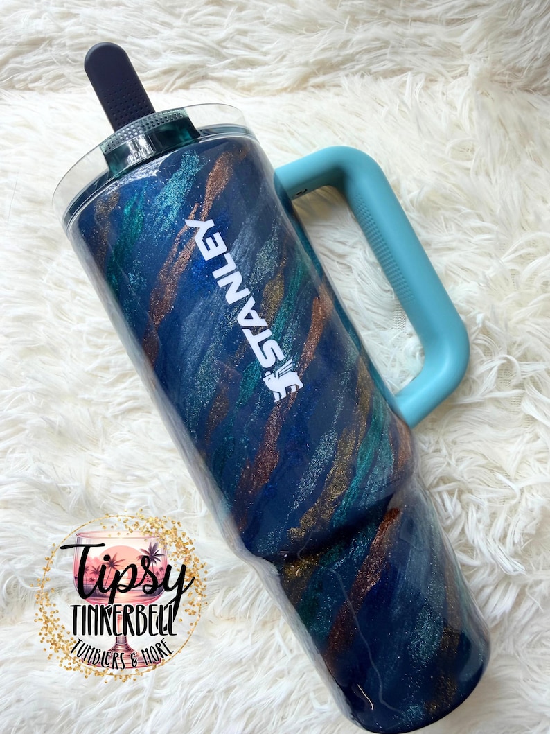 Puede incluir: Un vaso Stanley con un dise&ntilde;o de purpurina azul, verde azulado y dorado. El vaso tiene un asa azul claro y el logotipo de Stanley en blanco. Tiene una tapa azul oscuro y una pajita negra. Un recipiente para bebidas elegante.