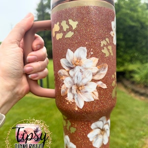 May include: A copper-coloured glitter tumbler with a handle and a dark translucent lid. The surface features white floral illustrations and gold-toned animal print accents. A circular logo with the text Tipsy Tinkerbell Tumblers & More is visible.