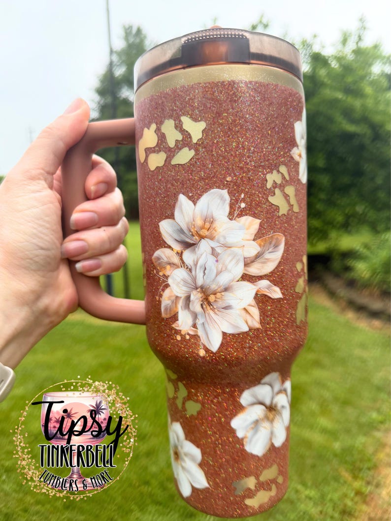 May include: A copper-coloured glitter tumbler with a handle and a dark translucent lid. The surface features white floral illustrations and gold-toned animal print accents. A circular logo with the text Tipsy Tinkerbell Tumblers & More is visible.