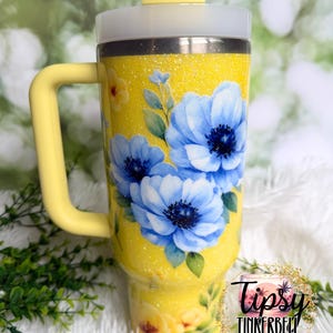 May include: A yellow glitter tumbler with a handle, featuring blue floral artwork. The tumbler has a white lid and a yellow straw. The text "Tipsy Tinkerbell Tumblers & More" is visible.
