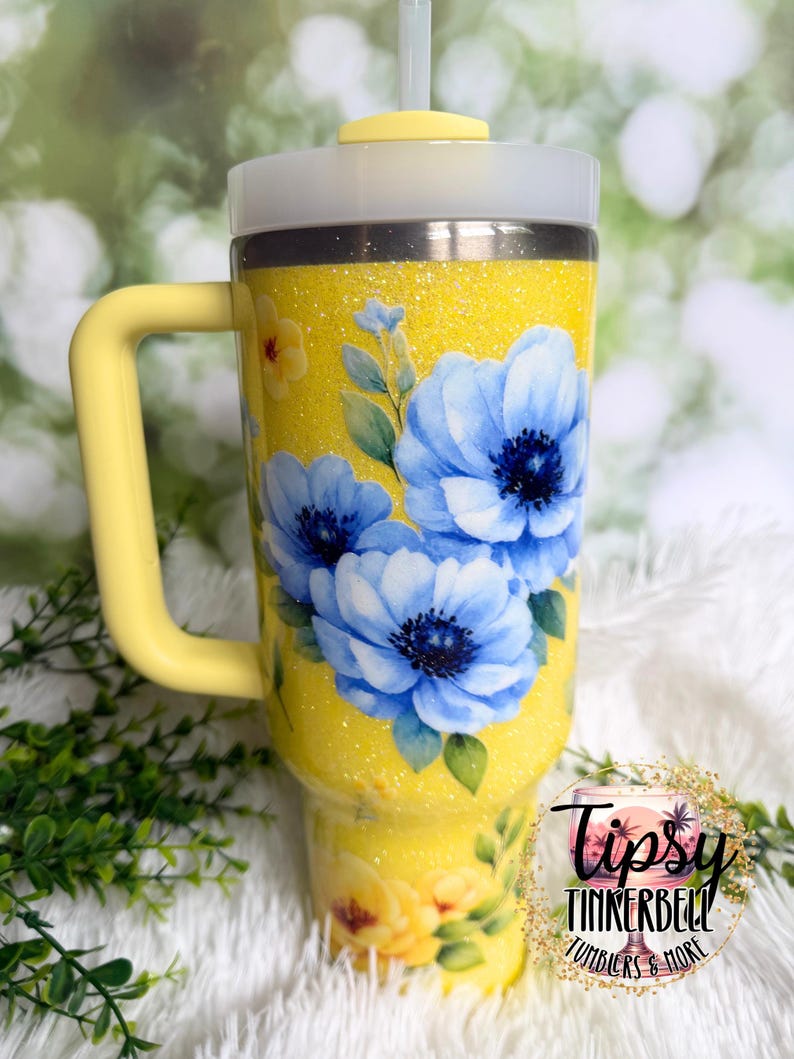 May include: A yellow glitter tumbler with a handle, featuring blue floral artwork. The tumbler has a white lid and a yellow straw. The text "Tipsy Tinkerbell Tumblers & More" is visible.