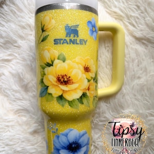May include: Yellow glitter Stanley tumbler with a floral design featuring yellow and blue flowers. The tumbler has a yellow handle and a white lid with a straw. The Stanley logo is visible on the front.