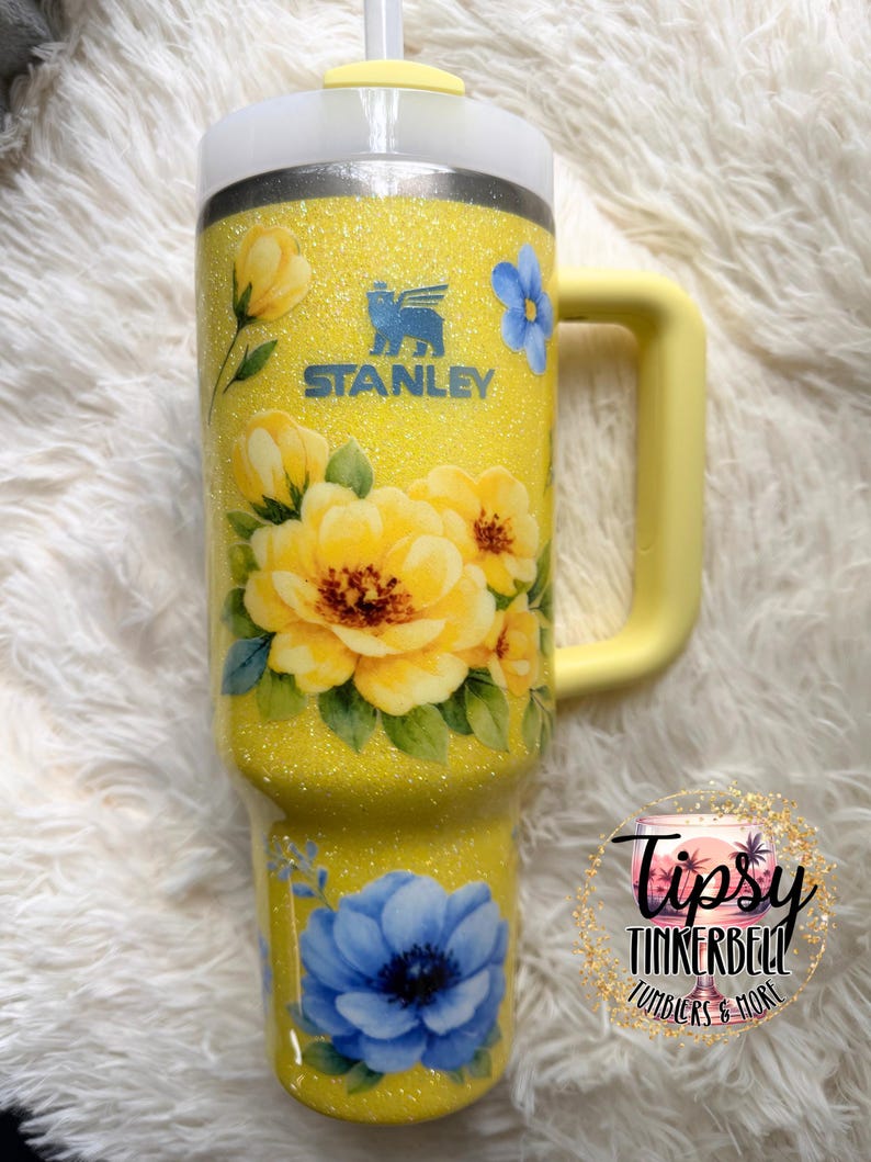 May include: Yellow glitter Stanley tumbler with a floral design featuring yellow and blue flowers. The tumbler has a yellow handle and a white lid with a straw. The Stanley logo is visible on the front.