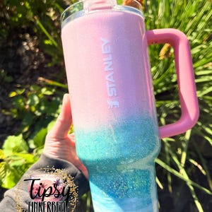 May include: A Stanley tumbler featuring a glittery ombre finish that transitions from light pink at the top to turquoise and blue in the middle, ending with a gold glitter base. It includes a pink handle, a clear lid, and a pink straw.