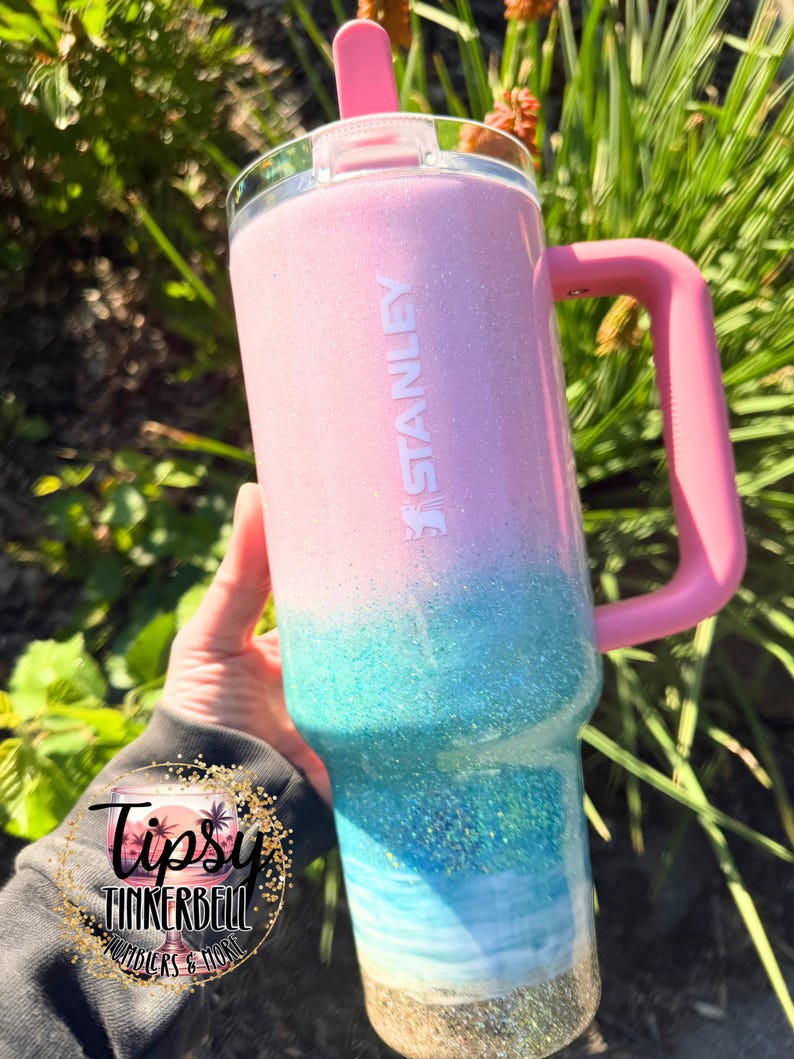 May include: A Stanley tumbler featuring a glittery ombre finish that transitions from light pink at the top to turquoise and blue in the middle, ending with a gold glitter base. It includes a pink handle, a clear lid, and a pink straw.