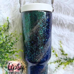 May include: A cylindrical stainless steel tumbler with a white lid and handle, featuring a dark, swirling glitter finish in shades of deep blue, emerald green, and black. The surface is coated in a glossy, reflective resin with embedded flakes.