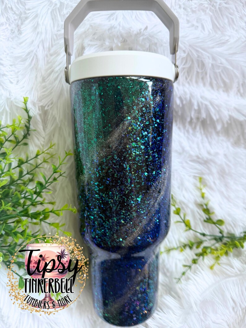 May include: A cylindrical stainless steel tumbler with a white lid and handle, featuring a dark, swirling glitter finish in shades of deep blue, emerald green, and black. The surface is coated in a glossy, reflective resin with embedded flakes.