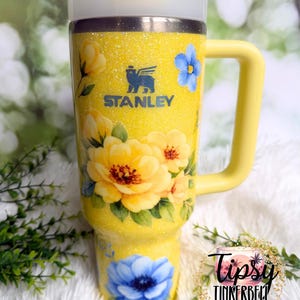 May include: A yellow Stanley tumbler with a floral design. The tumbler features a yellow handle, lid, and straw. The body of the tumbler is covered in yellow glitter and decorated with yellow and blue flowers. The Stanley logo is visible on the front.