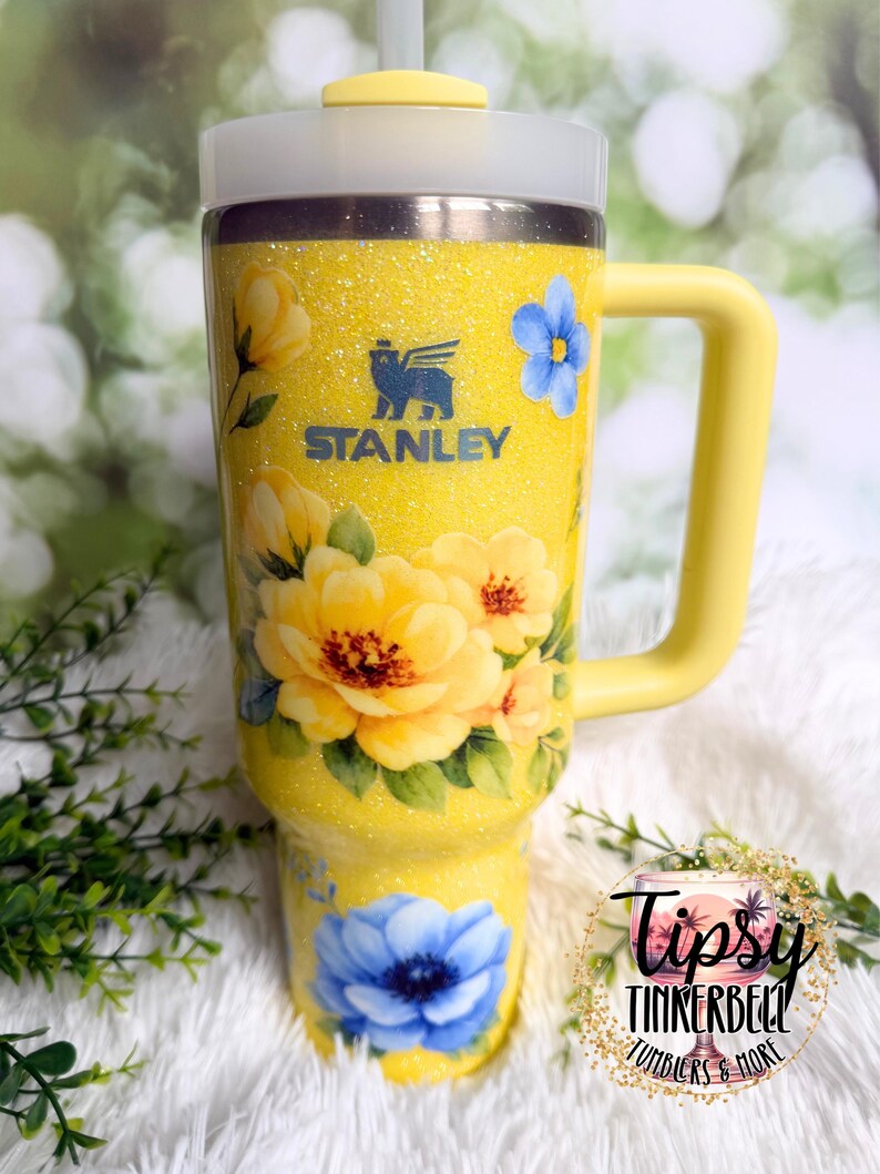 May include: A yellow Stanley tumbler with a floral design. The tumbler features a yellow handle, lid, and straw. The body of the tumbler is covered in yellow glitter and decorated with yellow and blue flowers. The Stanley logo is visible on the front.