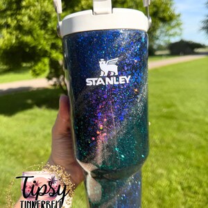 May include: A blue and green glitter tumbler with a white lid and handle. The Stanley logo is printed in white on the front. The surface features a swirling galaxy-like pattern of dark blue, black, and teal glitter with reflective highlights.