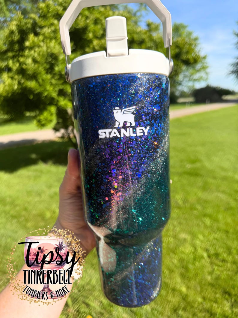 May include: A blue and green glitter tumbler with a white lid and handle. The Stanley logo is printed in white on the front. The surface features a swirling galaxy-like pattern of dark blue, black, and teal glitter with reflective highlights.
