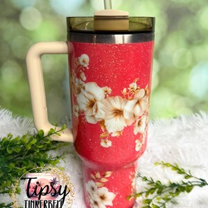 K&ouml;nnte beinhalten: Ein korallenfarbener Becher mit Glitzer-Finish, verziert mit einem Blumenmuster aus wei&szlig;en und cremefarbenen Bl&uuml;ten. Der Becher verf&uuml;gt &uuml;ber einen beigen Henkel, einen dunkel get&ouml;nten Deckel und einen transparenten Strohhalm. Ein kreisf&ouml;rmiges Logo zeigt Tipsy Tinkerbell Tumblers & More.