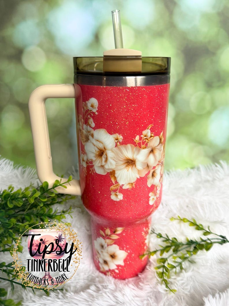 K&ouml;nnte beinhalten: Ein korallenfarbener Becher mit Glitzer-Finish, verziert mit einem Blumenmuster aus wei&szlig;en und cremefarbenen Bl&uuml;ten. Der Becher verf&uuml;gt &uuml;ber einen beigen Henkel, einen dunkel get&ouml;nten Deckel und einen transparenten Strohhalm. Ein kreisf&ouml;rmiges Logo zeigt Tipsy Tinkerbell Tumblers & More.