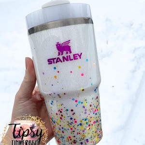 Puede incluir: Un vaso Stanley blanco con acabado brillante y detalles de confeti de colores. El vaso presenta el logotipo de Stanley en morado y una tapa blanca con pajita. La parte inferior del vaso est&aacute; cubierta de confeti de colores.