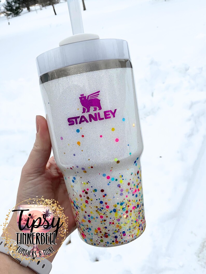 Puede incluir: Un vaso Stanley blanco con acabado brillante y detalles de confeti de colores. El vaso presenta el logotipo de Stanley en morado y una tapa blanca con pajita. La parte inferior del vaso est&aacute; cubierta de confeti de colores.