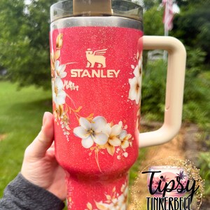 K&ouml;nnte beinhalten: Ein korallenfarbener Stanley-Becher mit Glitzer-Finish, verziert mit einem wei&szlig;en Blumenmuster und einem beigen Henkel. Der Becher hat einen transparenten Deckel mit beiger Lasche und einen Strohhalm. Ein kreisf&ouml;rmiges Logo tr&auml;gt den Text Tipsy Tinkerbell Tumblers & More.