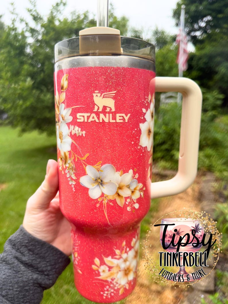 K&ouml;nnte beinhalten: Ein korallenfarbener Stanley-Becher mit Glitzer-Finish, verziert mit einem wei&szlig;en Blumenmuster und einem beigen Henkel. Der Becher hat einen transparenten Deckel mit beiger Lasche und einen Strohhalm. Ein kreisf&ouml;rmiges Logo tr&auml;gt den Text Tipsy Tinkerbell Tumblers & More.