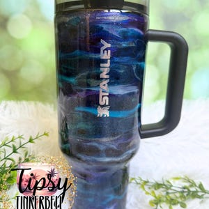 May include: A Stanley tumbler with a black handle and lid, featuring a swirling blue, purple, and silver design. The tumbler has the Stanley logo in white and the text "Tipsy Tinkerbell Tumblers & More" in a circular logo.
