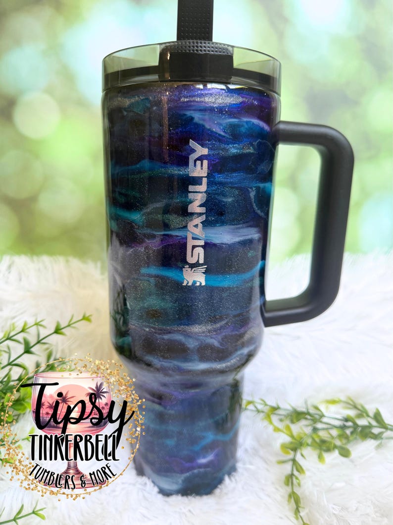 May include: A Stanley tumbler with a black handle and lid, featuring a swirling blue, purple, and silver design. The tumbler has the Stanley logo in white and the text "Tipsy Tinkerbell Tumblers & More" in a circular logo.