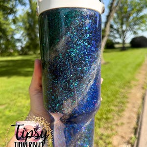 May include: A cylindrical tumbler with a white lid and handle, featuring a dark blue and black glitter finish with swirling silver accents. A circular logo on the lower left reads Tipsy Tinkerbell Tumblers & More in black script and print text.