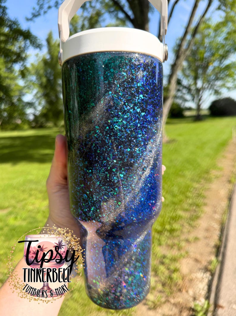 May include: A cylindrical tumbler with a white lid and handle, featuring a dark blue and black glitter finish with swirling silver accents. A circular logo on the lower left reads Tipsy Tinkerbell Tumblers & More in black script and print text.