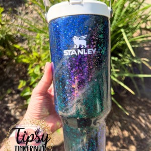 May include: A Stanley tumbler featuring a white lid and handle, with a dark blue, purple, and green glitter finish. The Stanley logo is printed in white on the upper portion of the tumbler. A circular logo with the text Tipsy Tinkerbell Tumblers & More is visible.