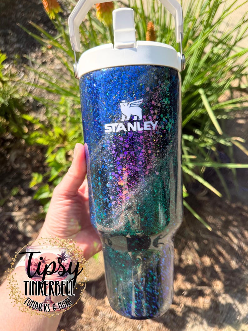 May include: A Stanley tumbler featuring a white lid and handle, with a dark blue, purple, and green glitter finish. The Stanley logo is printed in white on the upper portion of the tumbler. A circular logo with the text Tipsy Tinkerbell Tumblers & More is visible.