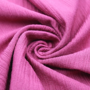 May include: Close-up of a swirl of magenta fabric. The fabric has a ribbed texture, with vertical lines running along its surface. The material is soft and appears to be knit or woven, with a slight sheen. The folds create a spiral pattern.